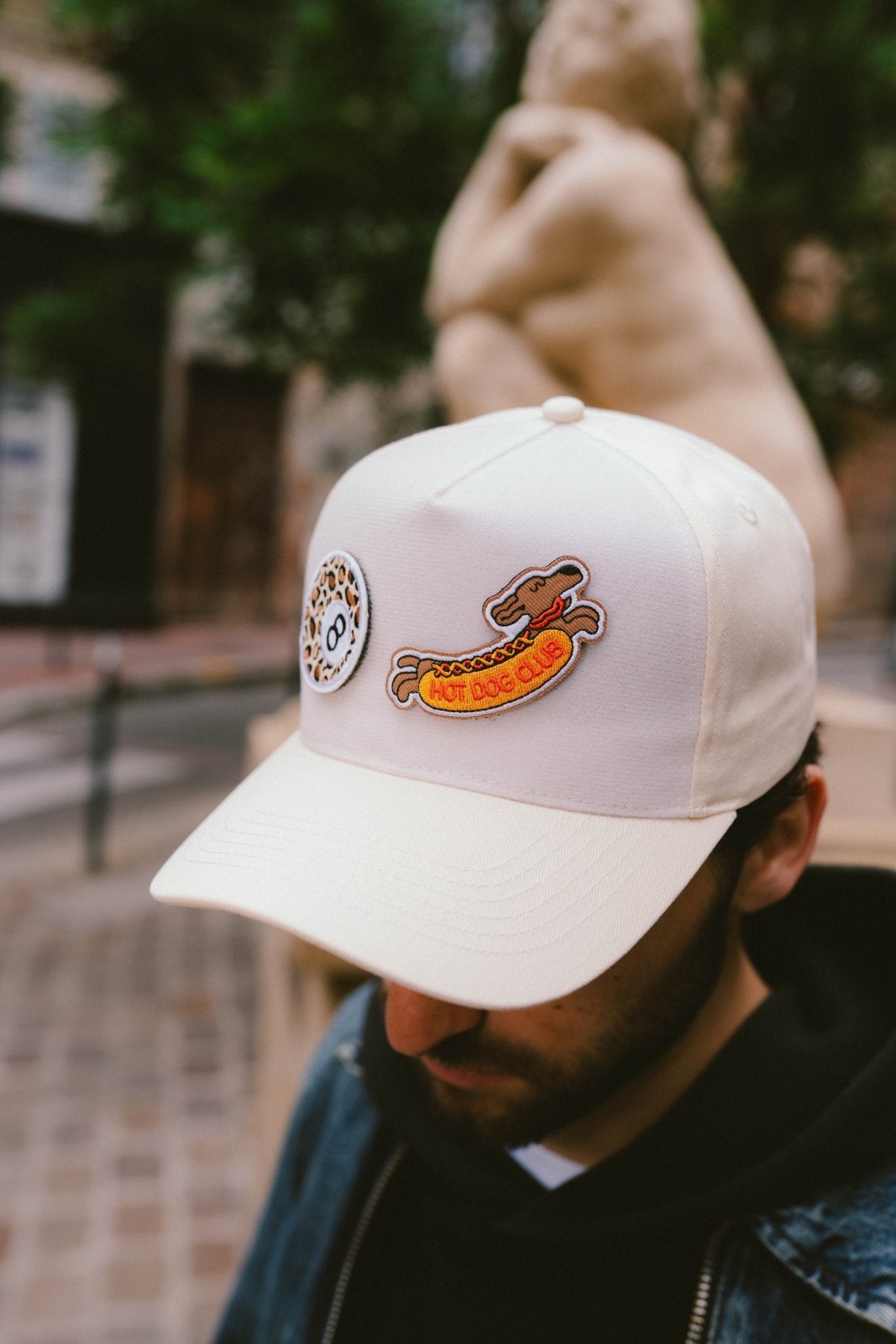 Gros plan sur une casquette Muzo beige personnalisée avec deux patchs scratchables : un teckel "Hot Dog Club" et une boule 8 motif léopard. Style streetwear urbain.