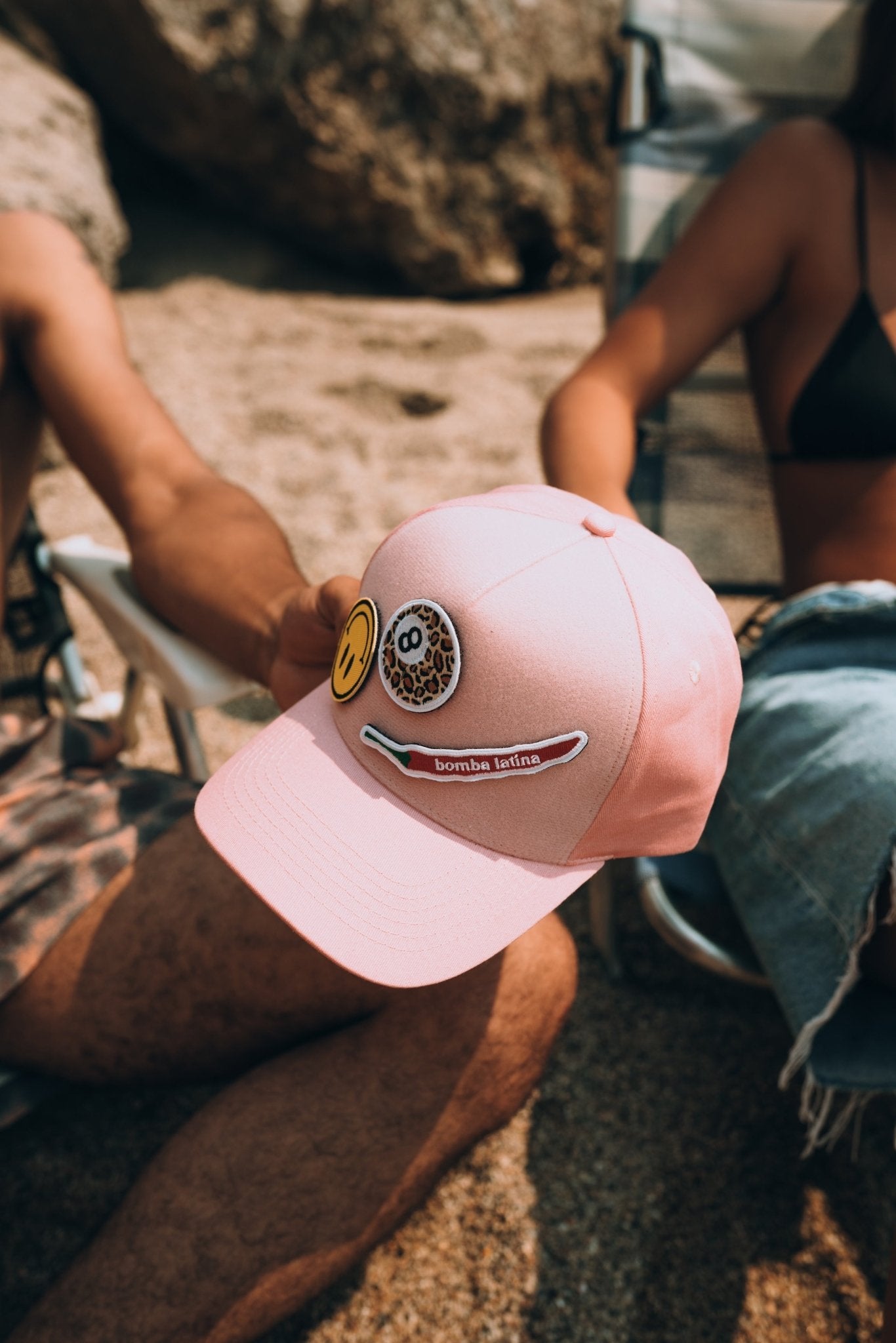 Casquette rose Muzo personnalisée avec patchs fun posée sur les genoux lors d'une journée à la plage, accessoire mode été.